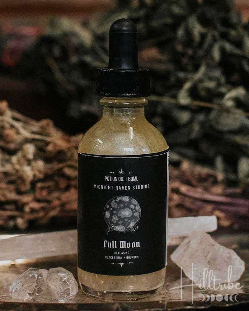 A small glass dropper bottle labelled 'Full Moon' sits on a rustic wooden surface with crystals, dried herbs, and a blurred wood background.