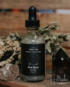 A glass dropper bottle labelled 'New Moon' with a black dropper cap, placed on a rustic wooden surface beside dried botanicals, a pink crystal, and a dark pointed crystal against a blurred wood background.