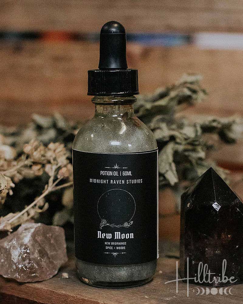 A glass dropper bottle labelled 'New Moon' with a black dropper cap, placed on a rustic wooden surface beside dried botanicals, a pink crystal, and a dark pointed crystal against a blurred wood background.