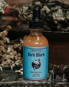 A bottle of 'Rich Witch' herbal tincture by Midnight Raven Studios featuring a light blue label and black dropper cap, set in a rustic environment with dried herbs and a glass dish.
