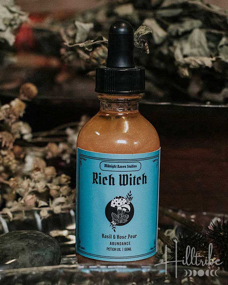 A bottle of 'Rich Witch' herbal tincture by Midnight Raven Studios featuring a light blue label and black dropper cap, set in a rustic environment with dried herbs and a glass dish.