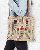 Person holding a crocheted bag with colorful handles against a white brick wall.