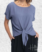 Woman wearing a blue top with a knot detail in front of a white brick wall, with 'HillBilly' branding.