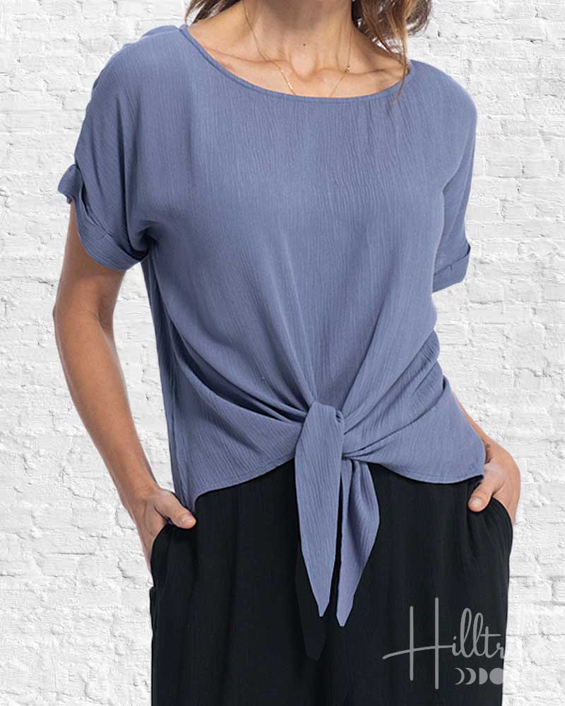 Woman wearing a blue top with a knot detail in front of a white brick wall, with 'HillBilly' branding.