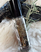 A clear glass roller bottle filled with dried botanicals, topped with a black cap, resting on a white faux-fur surface with dark wood and rosemary sprigs.