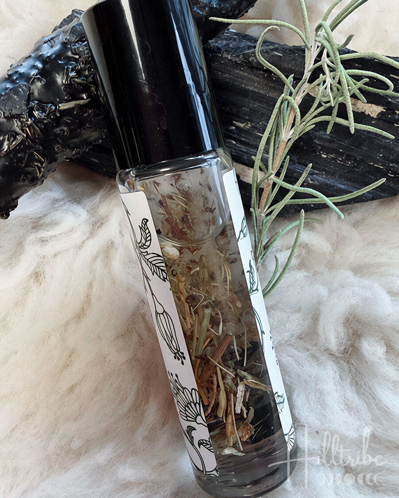 A clear glass roller bottle filled with dried botanicals, topped with a black cap, resting on a white faux-fur surface with dark wood and rosemary sprigs.