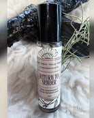 A 10ml glass roller bottle of 'Return to Sender' crystal infused rose oil, featuring a black cap and a white label with black text and illustrations. The bottle is styled on a white faux fur surface with a black wood piece and rosemary sprigs in the background.