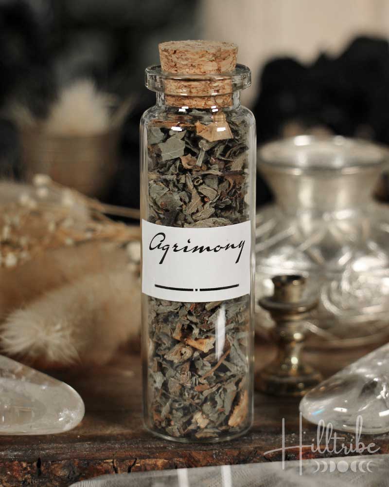 Glass bottle with cork lid containing dried herbs labeled 'Agrimony' on a wooden surface.