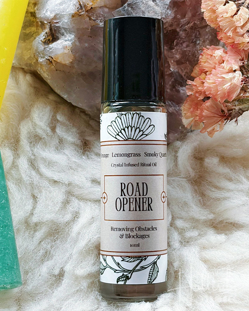 A 10ml bottle of Road Opener Lemongrass Stimule QUADE crystal infused blend oil, featuring a black cap and white label with black text, styled on a white faux fur surface with crystals and dried flowers.