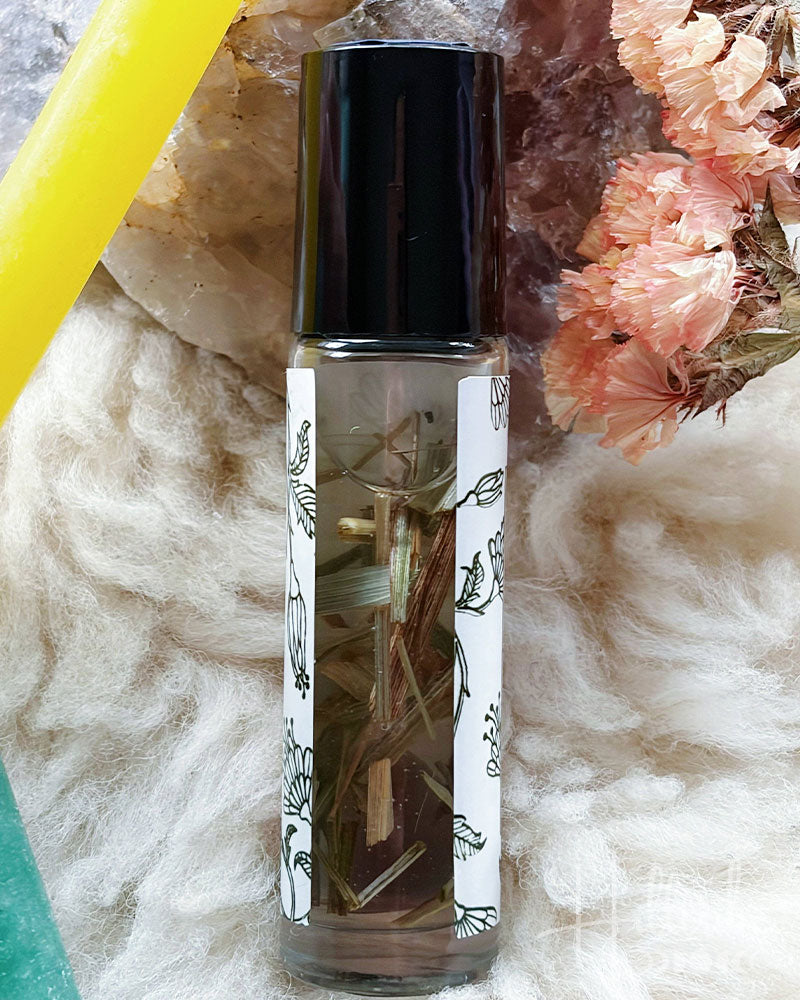 A clear glass roller bottle with a black cap and a white illustrated label, filled with botanical-infused oil, set against a soft white faux-fur background with crystals and dried flowers.