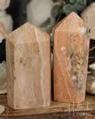 Close-up of two Rose Amethyst + Marble Jasper Tower on a wooden surface with a blurred background of decorative items.