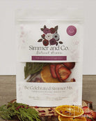 A stand-up pouch of Simmer and Co. Natural Aroma home fragrance, labeled 'Be Celebrated Simmer Mix,' with dried orange slices, strawberries, and greenery arranged around it on a tabletop.