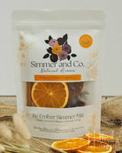 A stand-up pouch of Simmer and Co. Be Ember Simmer Mix natural home fragrance, featuring dried orange slices and a coconut visible through the window, styled on burlap with dried grass and orange slices in the foreground.