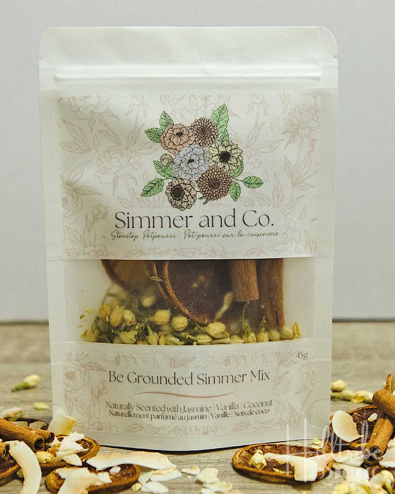A stand-up pouch of Simmer and Co. Be Grounded Simmer Mix herbal tea blend, shown on a tabletop with cinnamon sticks, coconut flakes, and tea leaves visible through the clear window.