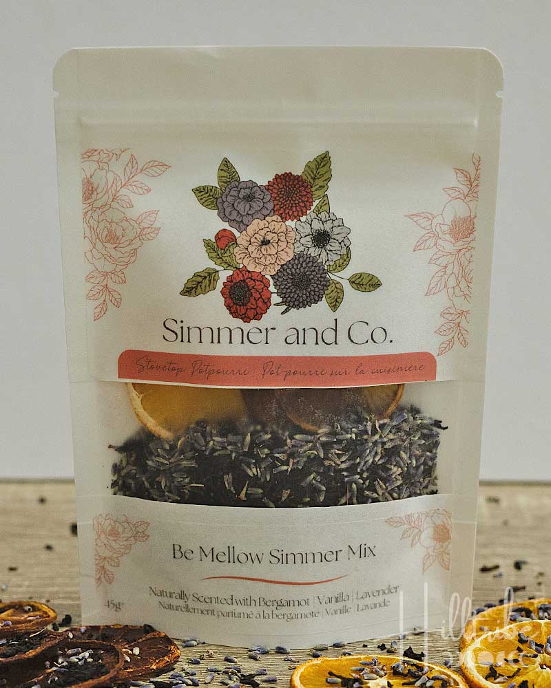 A stand-up pouch of Simmer and Co. Be Mellow Simmer Mix herbal tea, featuring a floral illustration and a clear window showing dried herbs and citrus slices inside, set on a rustic wooden surface with dried orange slices and lavender scattered around.