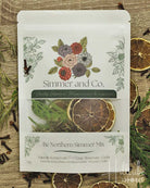 A stand-up pouch of Simmer and Co. 'Be Northern Simmer Mix' herbal tea, displayed on a rustic wooden surface surrounded by dried citrus slices, cloves, and rosemary sprigs.