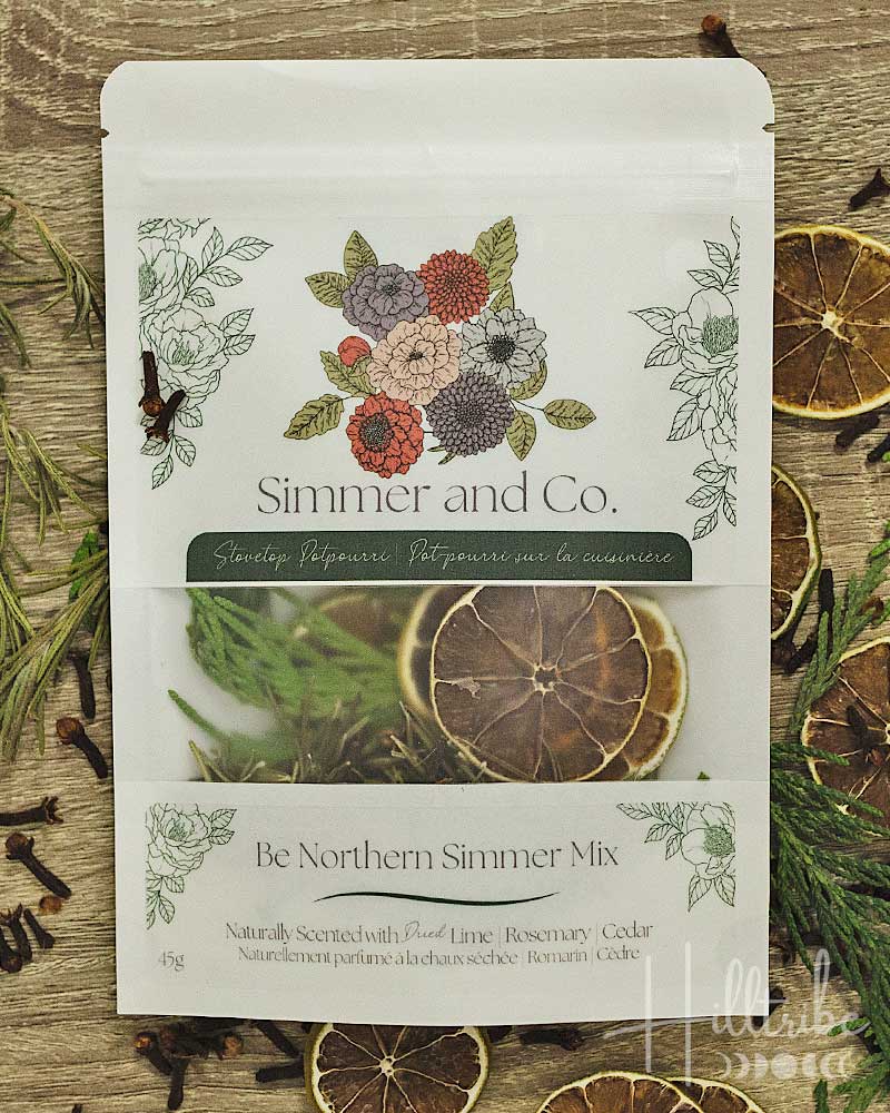 A stand-up pouch of Simmer and Co. 'Be Northern Simmer Mix' herbal tea, displayed on a rustic wooden surface surrounded by dried citrus slices, cloves, and rosemary sprigs.