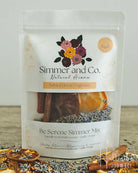 A stand-up pouch of Simmer and Co. Be Serene Simmer Mix natural home fragrance, showing dried lavender, cinnamon sticks, and citrus slices inside a clear window, with loose ingredients arranged around the base on a tabletop.