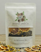 A stand-up pouch of Simmer and Co. Be Still Simmer Mix herbal tea, featuring dried citrus slices and lavender flowers visible through a clear window, with dried citrus slices scattered around on a tabletop.