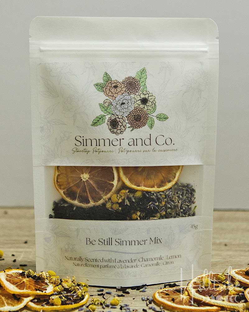 A stand-up pouch of Simmer and Co. Be Still Simmer Mix herbal tea, featuring dried citrus slices and lavender flowers visible through a clear window, with dried citrus slices scattered around on a tabletop.