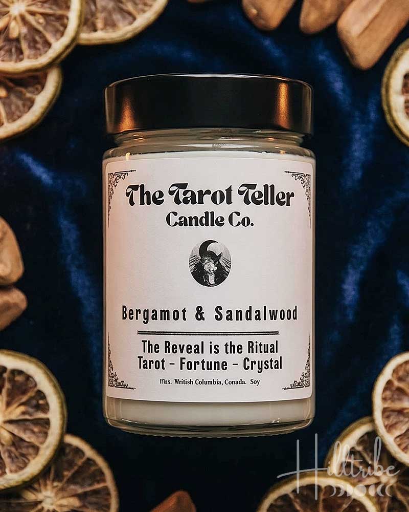 A jar candle labeled 'The Tarot Teller Candle Co.' in the scent Bergamot & Sandalwood, styled in a flat lay with dried orange slices, sandalwood pieces, a pocket watch, and tarot cards on a dark blue velvet background.
