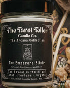 A jar candle labeled 'The Emperors Elixir' by The Tarot Teller Candle Co. is centered over an arranged flat lay of tarot cards, crystals, dried leaves, and wax melts under warm candlelight.