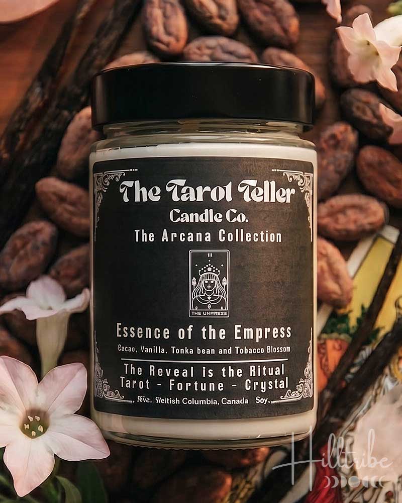 A jar candle labeled 'The Tarot Teller Candle Co. The Aroha Collection' with the scent name 'Essence of the Empress,' styled on a wooden surface with cacao beans, vanilla beans, pink flowers, crystals, and tarot cards.