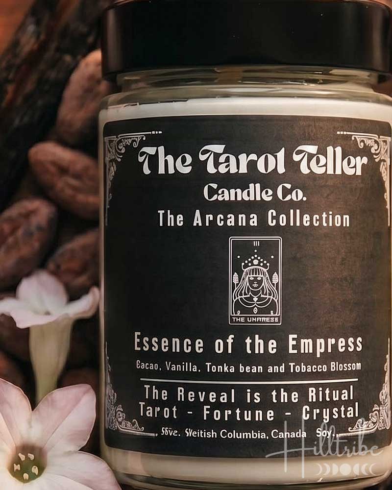A jar candle labeled 'The Tarot Teller Candle Co. The Aroha Collection' with the scent name 'Essence of the Empress,' styled on a wooden surface with cacao beans, vanilla beans, pink flowers, crystals, and tarot cards.