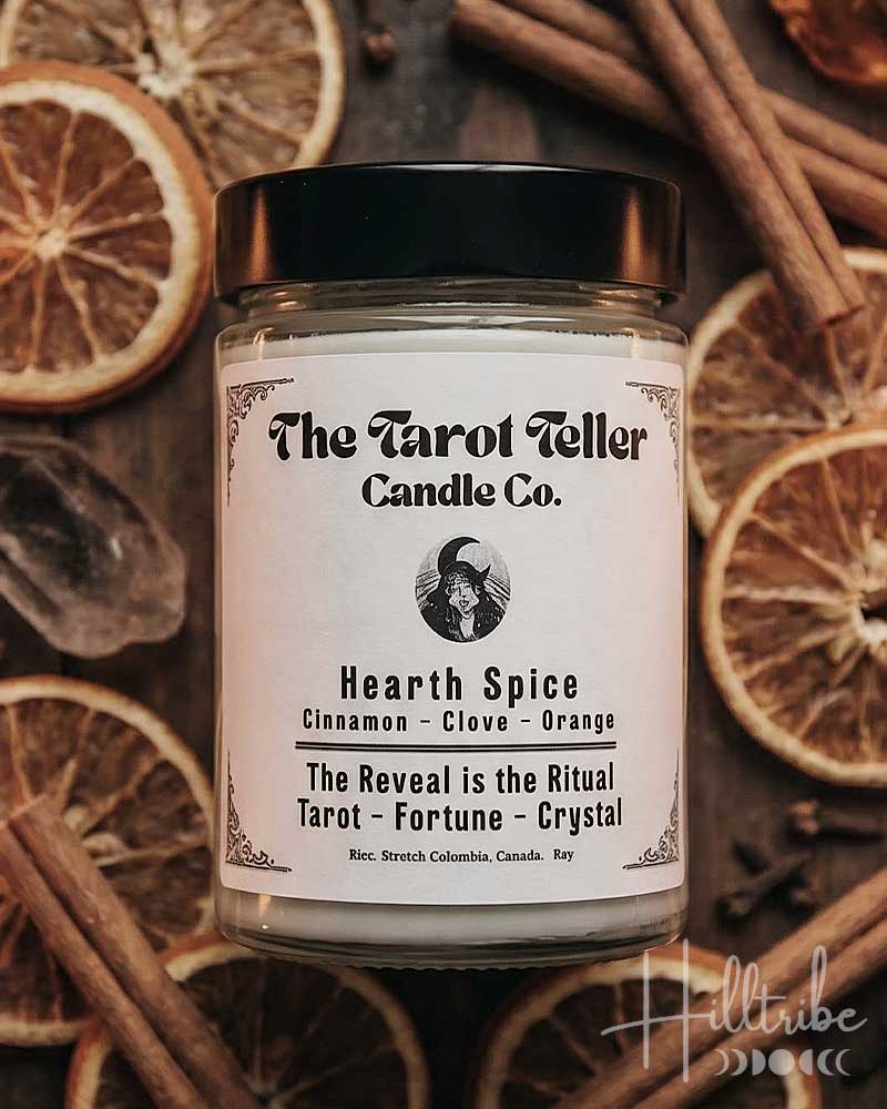 A jar candle labeled 'Hearth Spice' by The Tarot Teller Candle Co. is centered on a dark wooden surface surrounded by dried orange slices, cinnamon sticks, cloves, and crystals.