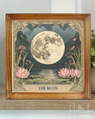A framed art print titled 'THE MOON' showing a large detailed full moon over a pond with pink lotus flowers, displayed on a wooden shelf next to a potted green plant.