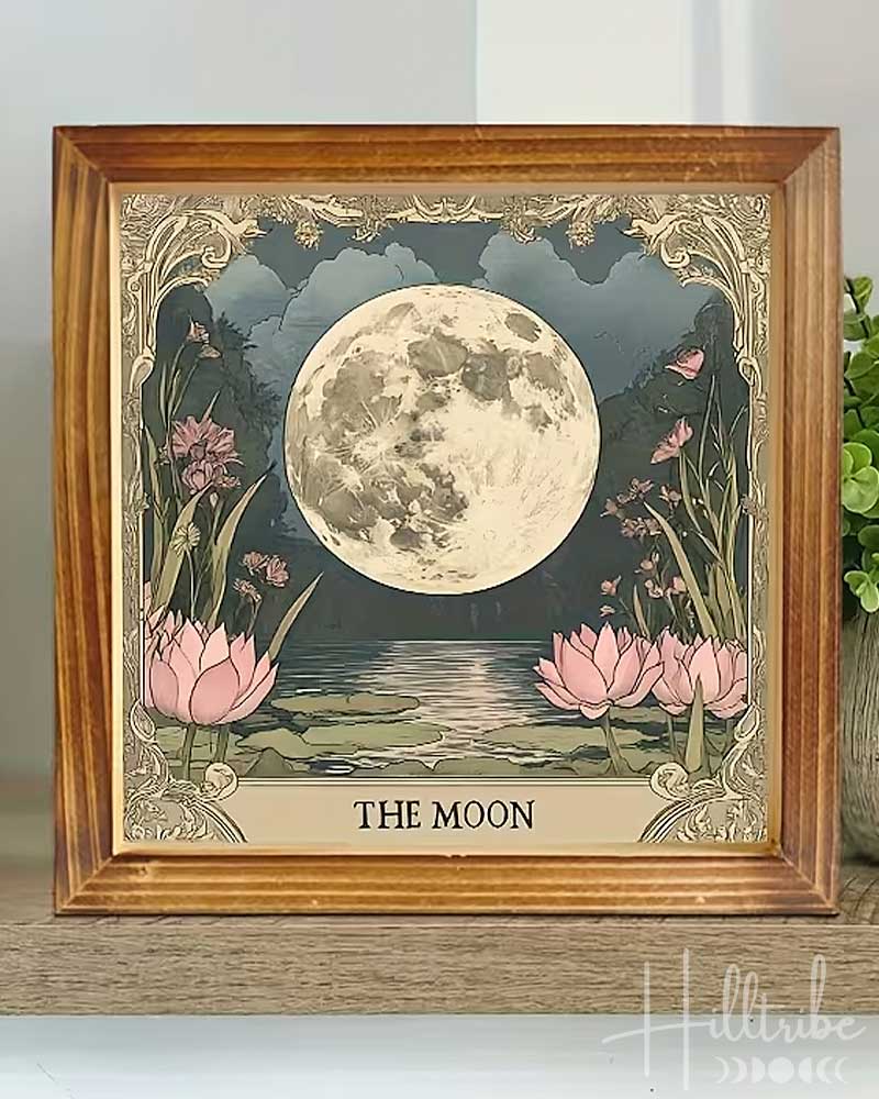 A framed art print titled 'THE MOON' showing a large detailed full moon over a pond with pink lotus flowers, displayed on a wooden shelf next to a potted green plant.