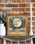 A framed art print titled 'THE MOON' featuring a detailed illustration of a full moon over pink water lilies, styled on a rustic wooden mantel with a white vase of greenery, white pillar candles, and a beaded tassel garland against a red brick wall.
