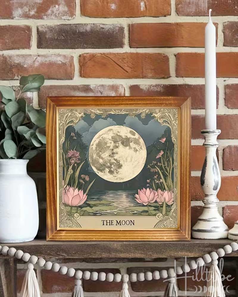 A framed art print titled 'THE MOON' featuring a detailed illustration of a full moon over pink water lilies, styled on a rustic wooden mantel with a white vase of greenery, white pillar candles, and a beaded tassel garland against a red brick wall.