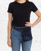 Person wearing a black t-shirt and blue jeans with a navy blue waist bag against a white brick wall.