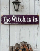 A rectangular wooden sign with a dark purple background and white lettering that reads “The Witch is in,” displayed against a white paneled door. The scene includes Halloween props: three small jack-o'-lanterns, a broom, bat cutouts, and a “2D FLAT PRINTING” label in the top left corner.