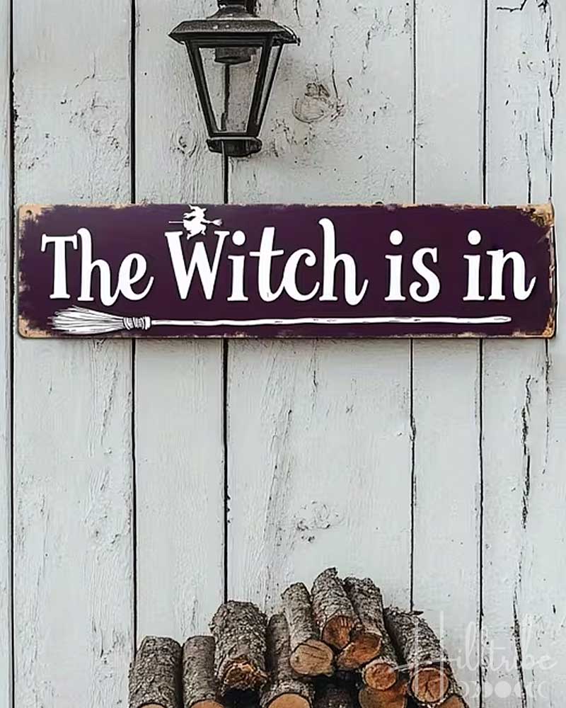 A rectangular wooden sign with a dark purple background and white lettering that reads “The Witch is in,” displayed against a white paneled door. The scene includes Halloween props: three small jack-o'-lanterns, a broom, bat cutouts, and a “2D FLAT PRINTING” label in the top left corner.