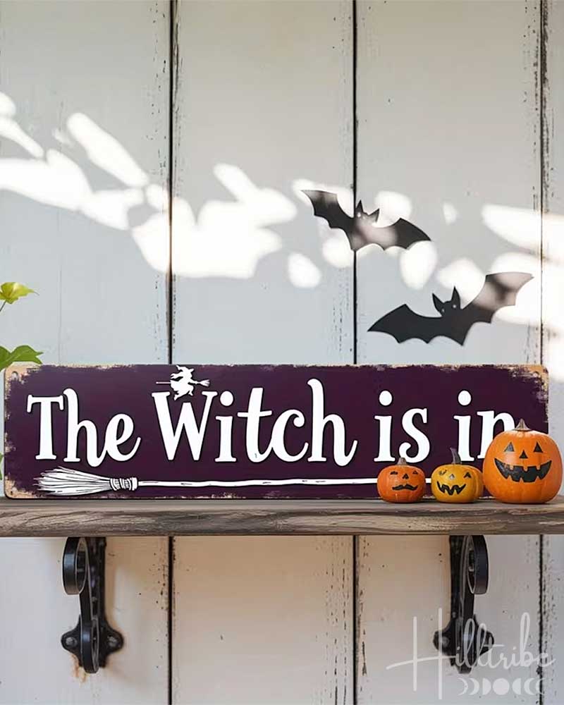 A rectangular wooden sign with a dark purple background and white lettering that reads “The Witch is in,” displayed on a rustic shelf against a white paneled door. The scene includes Halloween props: three small jack-o'-lanterns, a broom, bat cutouts, and a “2D FLAT PRINTING” label in the top left corner.