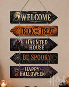 A 5-piece wooden arrow sign set for Halloween, hanging on a beige wall above a decorated mantel with pumpkins and candles. The sign features rustic distressed wood textures in black, orange, and purple, with cream lettering reading 'WELCOME', 'TRICK OR TREAT', 'HAUNTED HOUSE', 'BE SPOOKY', and 'HAPPY HALLOWEEN'.