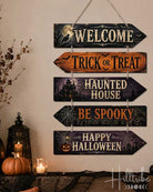 A 5-piece wooden arrow sign set for Halloween, hanging on a beige wall above a decorated mantel with pumpkins and candles. The sign features rustic distressed wood textures in black, orange, and purple, with cream lettering reading 'WELCOME', 'TRICK OR TREAT', 'HAUNTED HOUSE', 'BE SPOOKY', and 'HAPPY HALLOWEEN'.