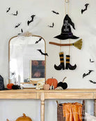 Halloween wall decor set featuring a witch hat garland with a smiling face, striped socks, a broom, and black bat cutouts, displayed on a white wall above a wooden console table with pumpkins and a mirror.