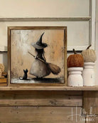 A framed flat wall art print showing a witch in a wide-brimmed hat sweeping a small black cat, displayed on a wooden shelf 