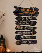 A rustic multi-tier wooden hanging sign with Halloween-themed text, displayed on a wall above a decorated table with pumpkins, candles, and dried branches. The sign reads: 'Witch's Halloween Rules' with additional phrases below.