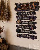 A rustic wooden hanging sign with the text 'Witch's Halloween Rules' and seven listed phrases, displayed on a cream wall in a styled room with pumpkins, dried flowers, and shelves.