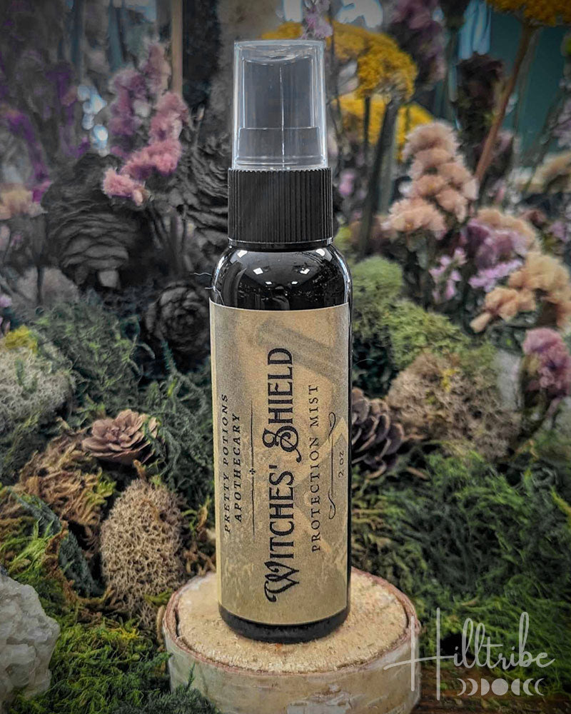 A small dark bottle labeled 'Witches' Shield Protection Mist' by Pretty Potions Apothecary, set against a natural background of moss, pinecones, and wood.
