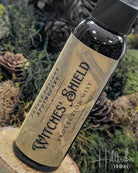 A small dark bottle labeled 'Witches' Shield Protection Mist' by Pretty Potions Apothecary, set against a natural background of moss, pinecones, and wood.
