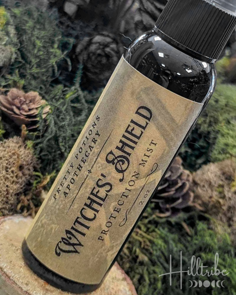 A small dark bottle labeled 'Witches' Shield Protection Mist' by Pretty Potions Apothecary, set against a natural background of moss, pinecones, and wood.